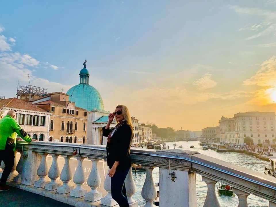 A woman stands on a bridge overlooking a canal in Venice, with a large turquoise-domed building in the background.