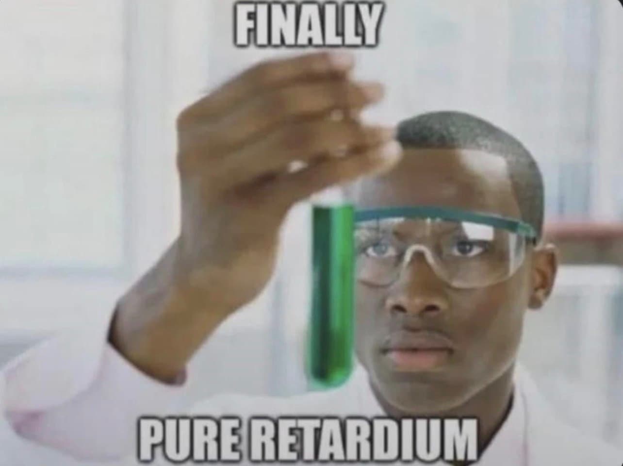 A man in a lab coat and safety goggles holds up a test tube with green liquid.