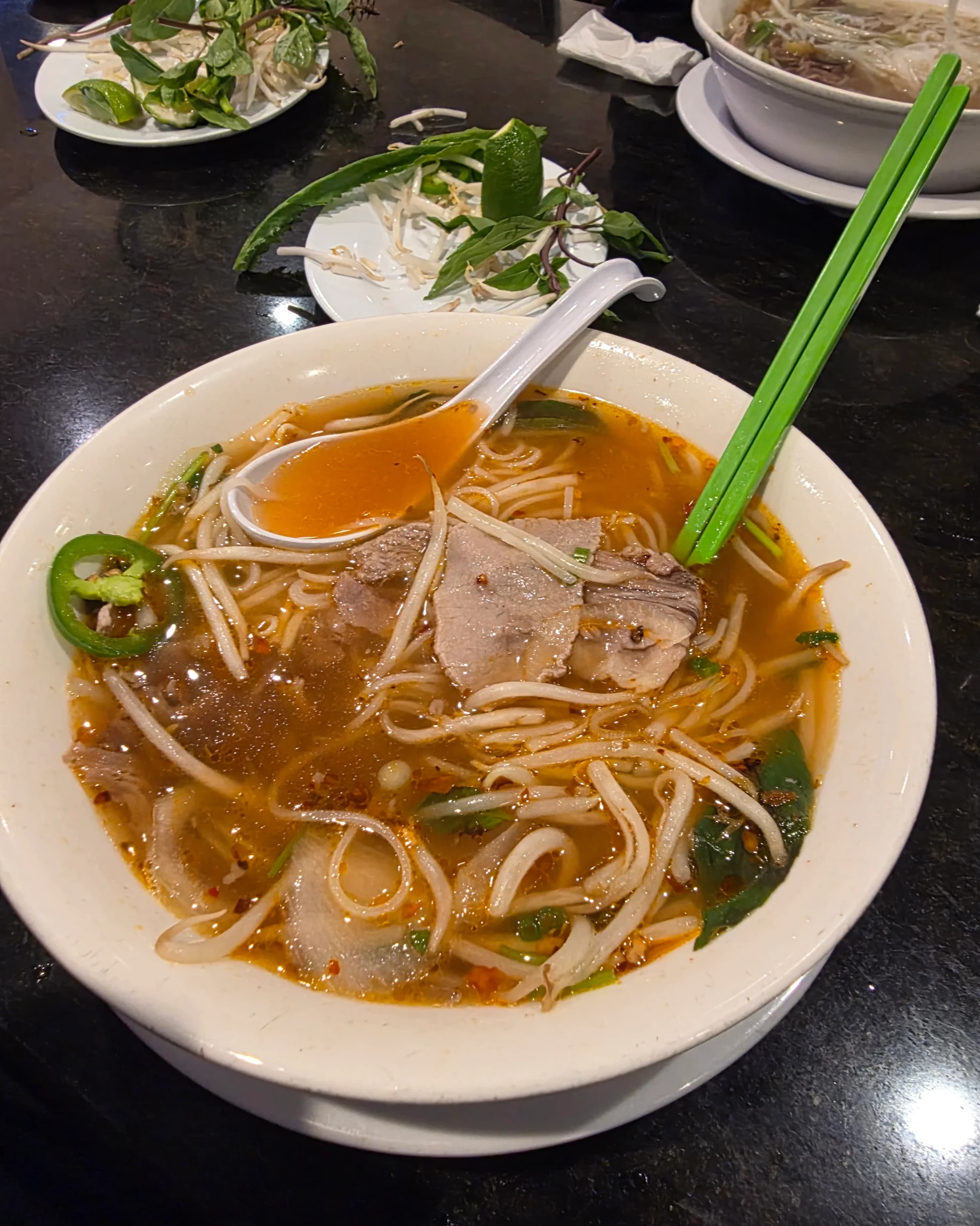 A bowl of pho with sliced meat, bean sprouts, and herbs in a spicy broth, accompanied by a spoon and chopsticks.