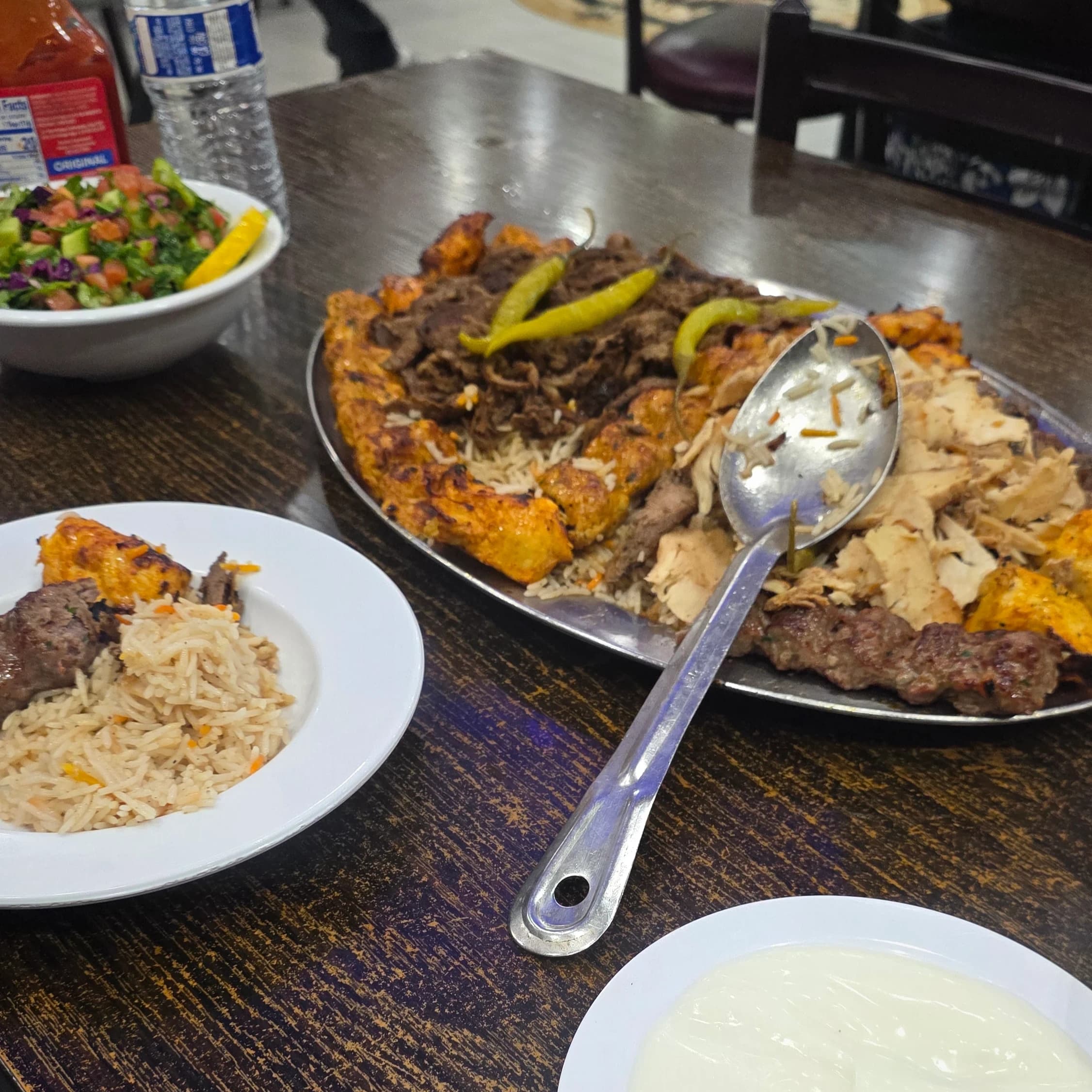 AI description: A large platter of grilled meats over rice, topped with green peppers and a serving spoon, sits on a restaurant table beside a plate of rice and meat, a chopped salad, and a bowl of white sauce.