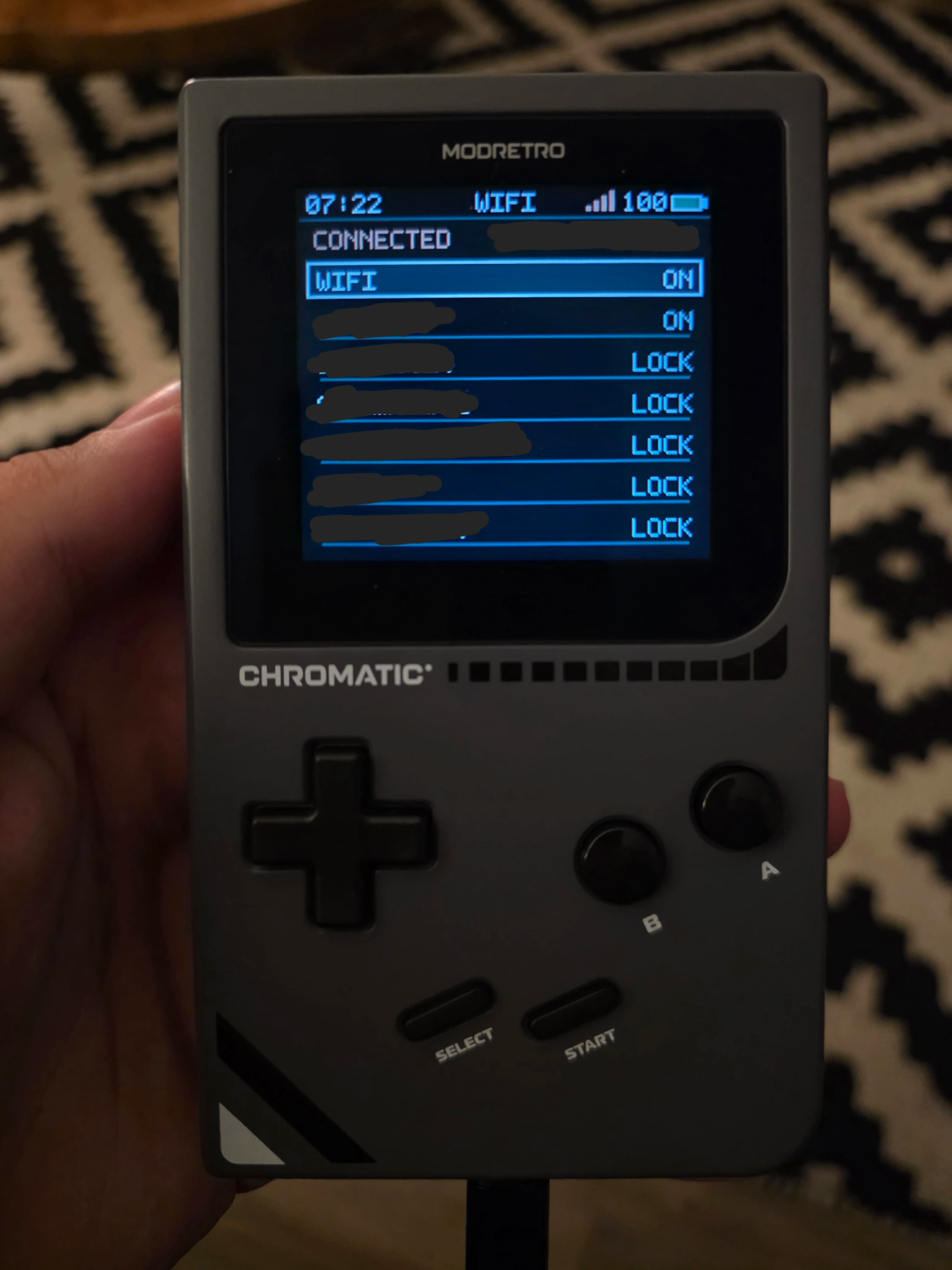 AI description: A hand holds a gray MODRETRO CHROMATIC handheld game console. Its screen shows a Wi-Fi menu with “CONNECTED,” “WIFI ON,” and several locked networks.