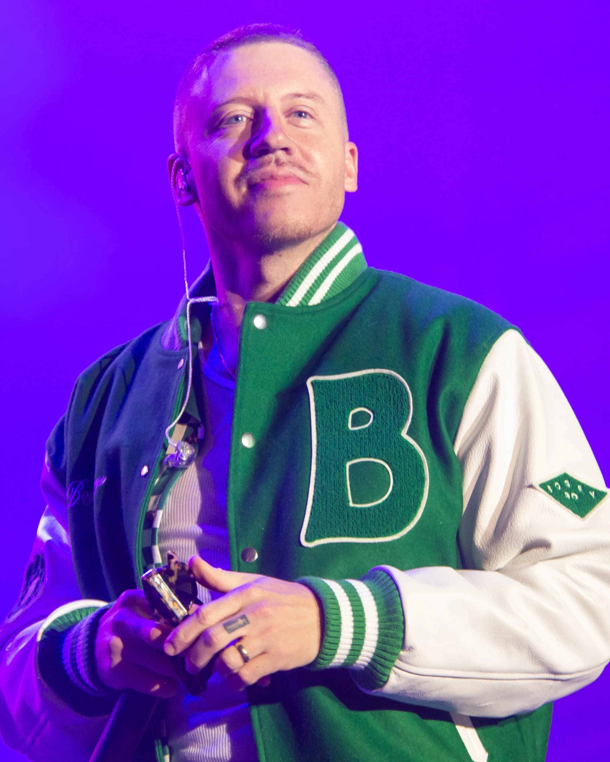 Singer Macklemore wearing a green and white varsity jacket with a large 'B' on the front, holding a phone.