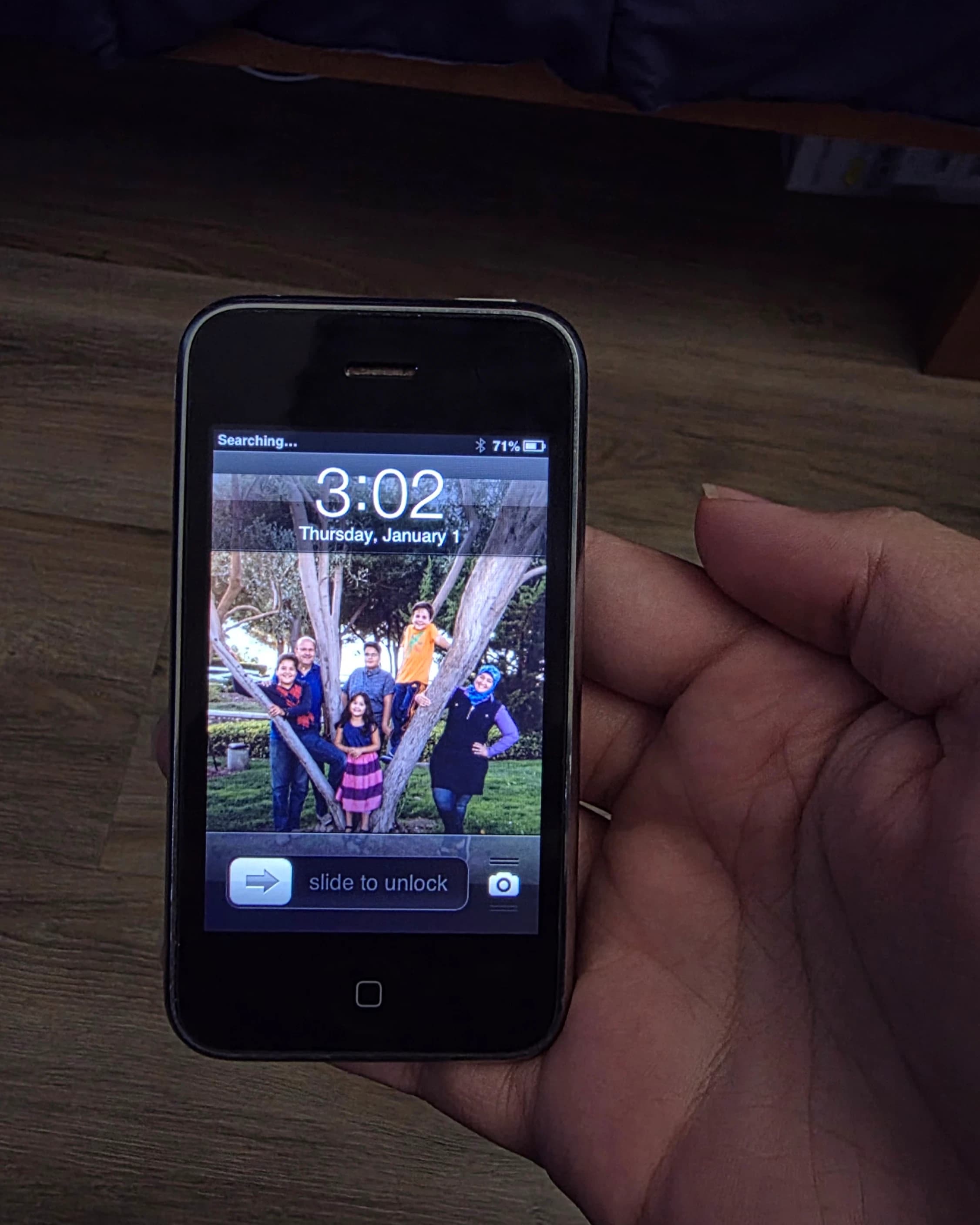 AI description: A hand holds an older black iPhone showing a lock screen with a group photo as its wallpaper. The screen reads “3:02,” “Thursday, January 1,” and “slide to unlock.”