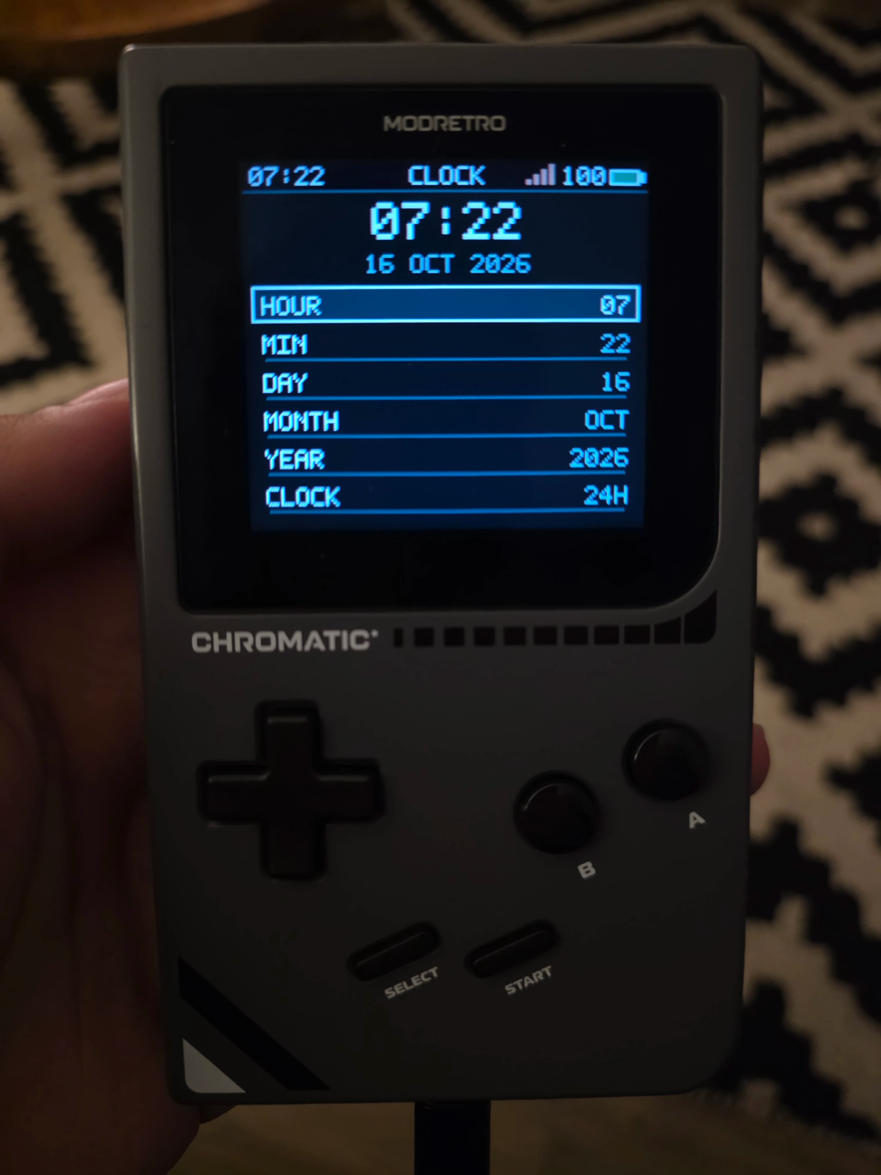 AI description: A hand holds a gray MODRETRO CHROMATIC handheld game console. Its blue-on-black clock screen shows “07:22” and “16 OCT 2026,” with hour, minute, day, month and year settings below.