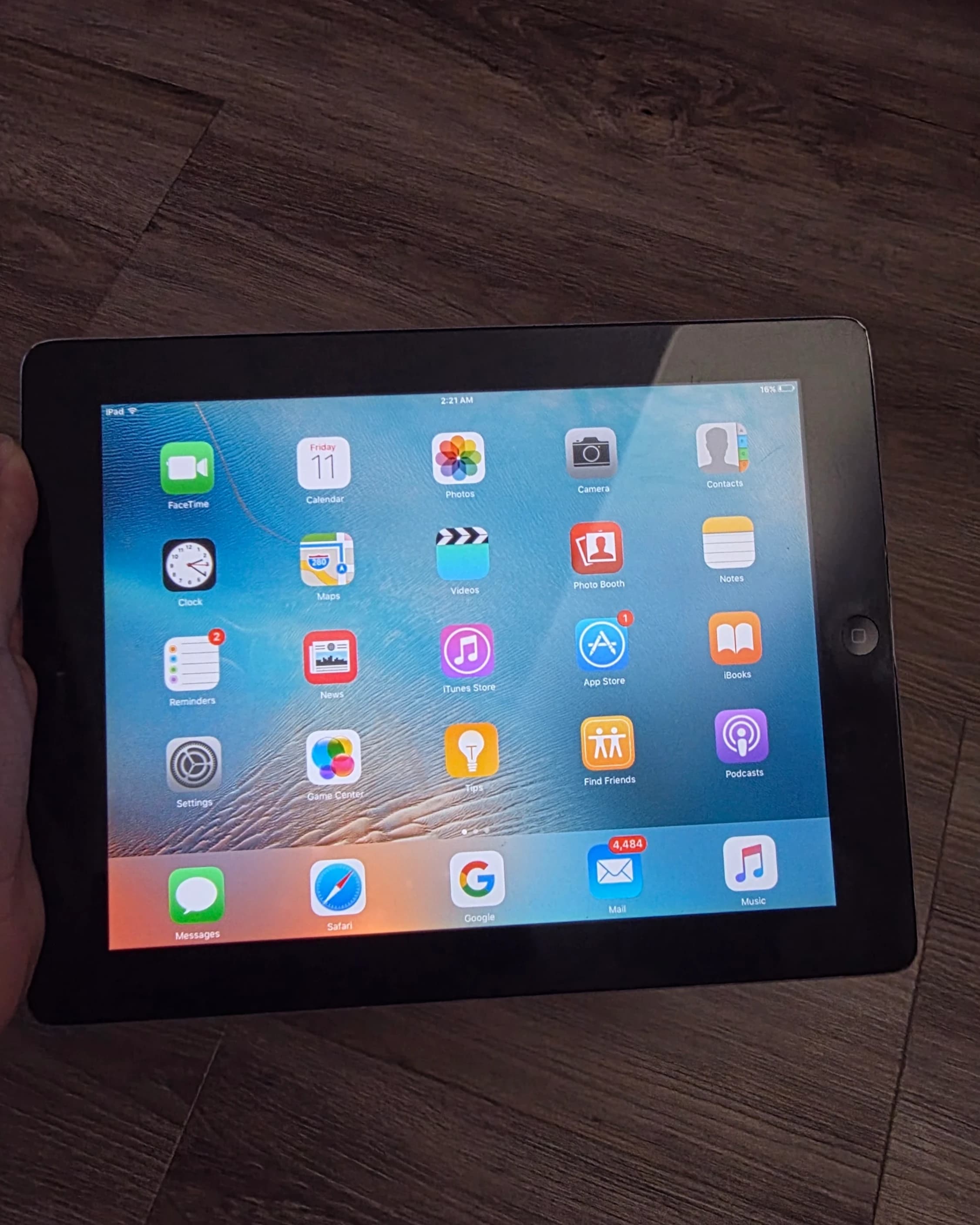 AI description: A black iPad rests on a wooden surface, displaying a home screen with app icons including FaceTime, Photos, Safari and Mail.