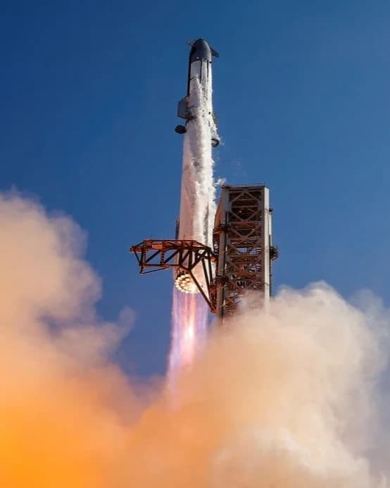 AI description: A tall silver rocket lifts off beside a launch tower, its engines glowing beneath it as thick orange-lit smoke billows across the foreground.