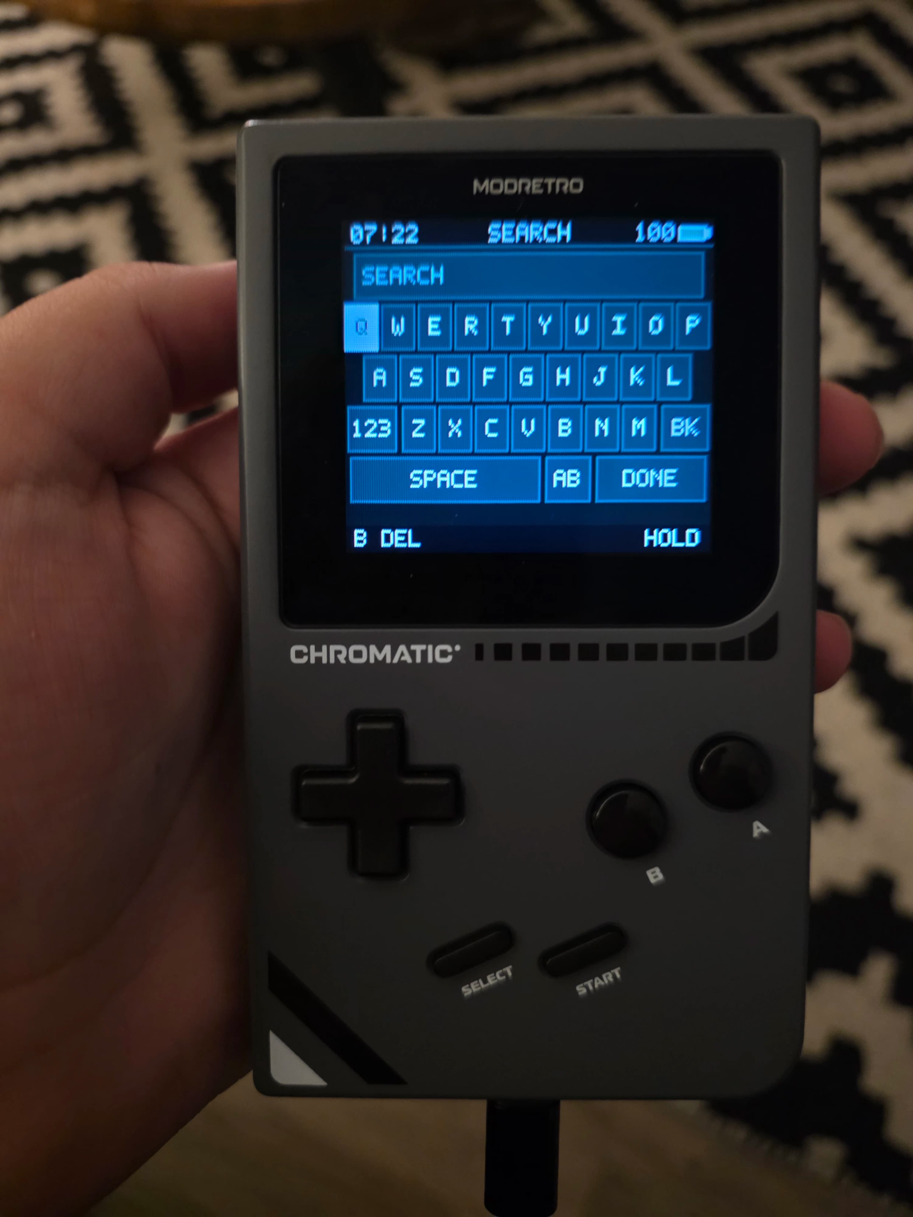 AI description: A hand holds a dark gray retro handheld game console with a blue keyboard screen showing “MODRETRO,” “SEARCH,” and “CHROMATIC”; a blurred pixel-art pattern is behind it.