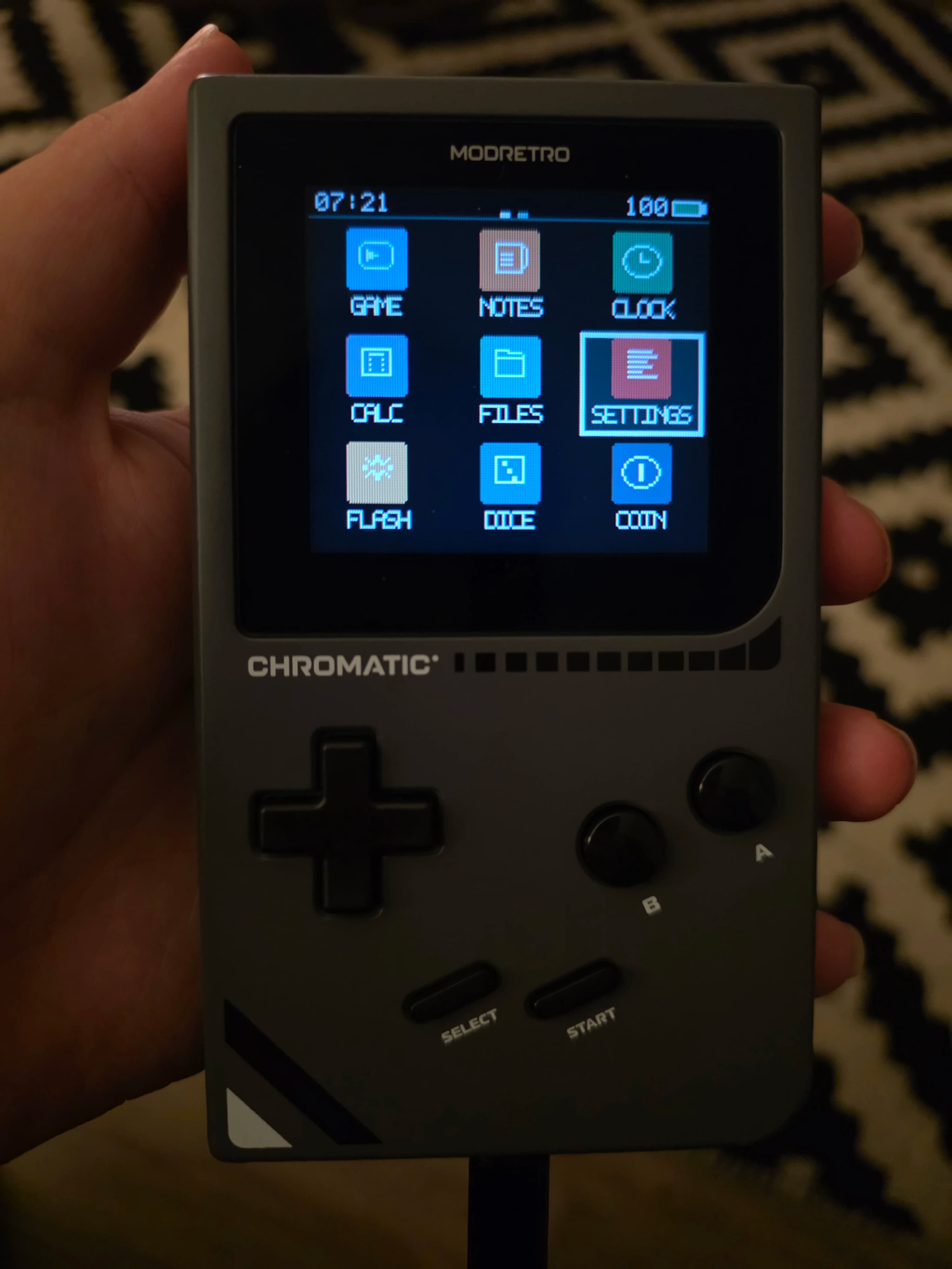 AI description: A hand holds a gray MODRETRO CHROMATIC handheld game console. Its screen shows a menu of apps, with “SETTINGS” selected.