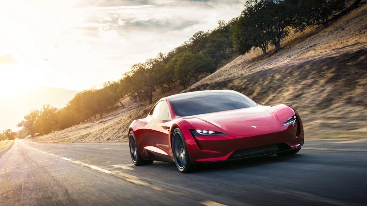 A red Tesla sports car driving on a road with a hillside and trees in the background.