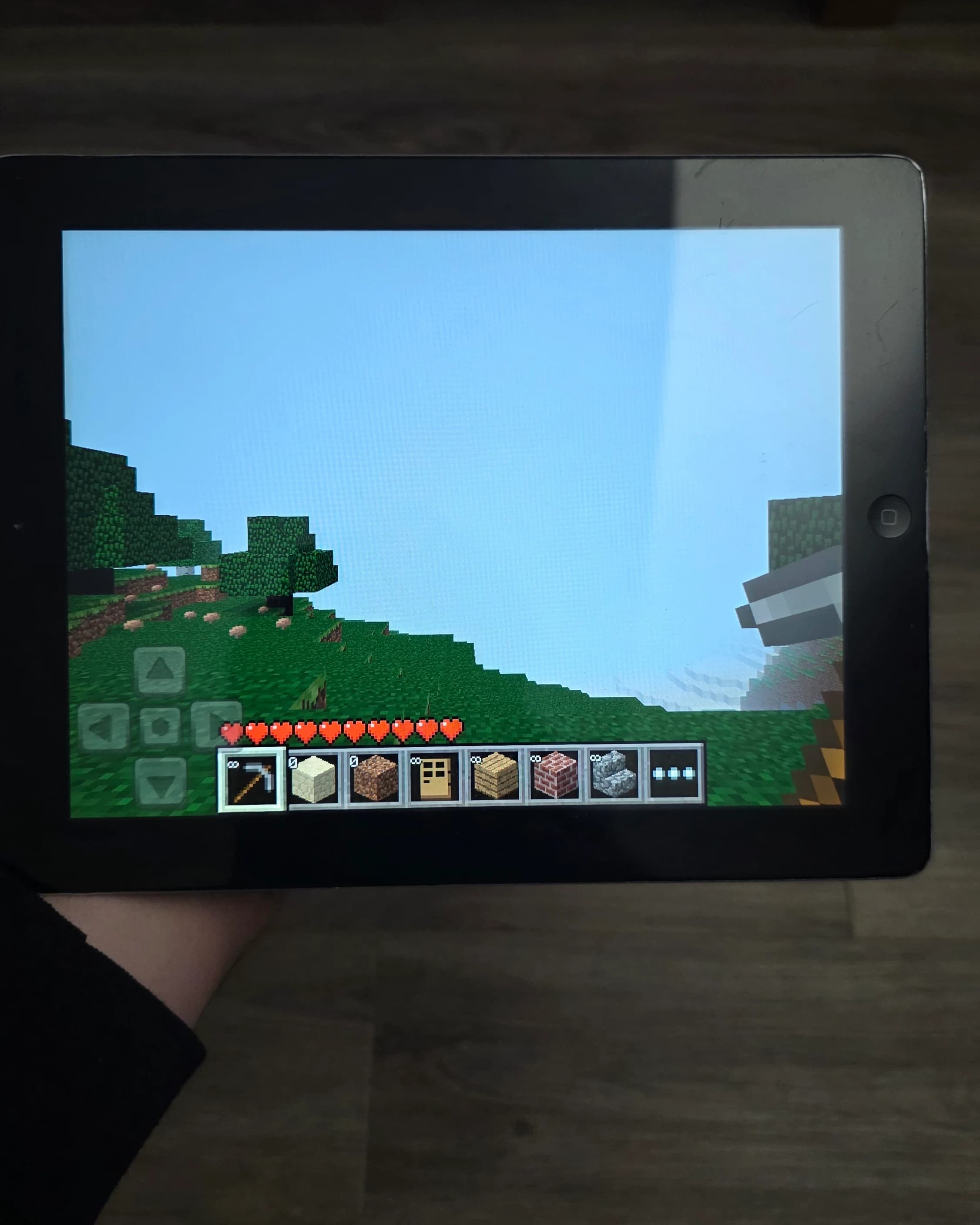 AI description: A black iPad held in one hand displays Minecraft, with blocky trees, grassy terrain and game controls on screen.